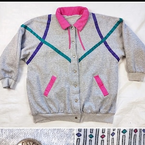 May include: A gray sweatshirt jacket with a pink, purple, and teal geometric design. The jacket has a snap closure and a pink collar. The jacket has a label that says "Current Seen".