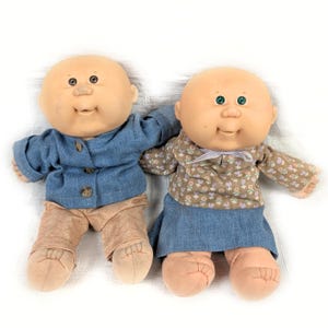 May include: Two vintage Cabbage Patch Kids dolls. One doll wears a blue denim jacket and tan pants. The other doll wears a floral print top and a blue denim skirt. Both dolls have bald heads, rosy cheeks, and smiling faces.