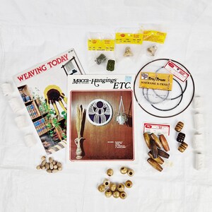 Puede incluir: Una colección de suministros de macramé que incluye cuentas de madera, cordón y un libro titulado "Macra-Hangings ETC.". El libro presenta una imagen colorida de una suspensión de macramé. El libro está rodeado de varios suministros de macramé, que incluyen cuentas de madera, cordón y un libro titulado "Macra-Hangings ETC."