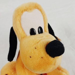 May include: A plush toy of Pluto, a cartoon dog from Disney. Pluto is a yellow and brown color with black ears and a black nose. He has large, black eyes.