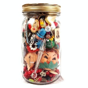 Op de afbeelding: Een doorzichtige glazen weckpot met een gouden metalen deksel, gevuld met een verzameling diverse kleine voorwerpen, waaronder een poppengezicht, rode kralen, een blauw plastic figuurtje, een witte knoop, een kleine vintage foto en een pipet.