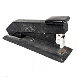 May include: A vintage black metal stapler with a textured, crinkle-finish body and a smooth, glossy black top press plate. The device features a silver-toned metal staple magazine at the front and a rectangular base with a small metal fastener.