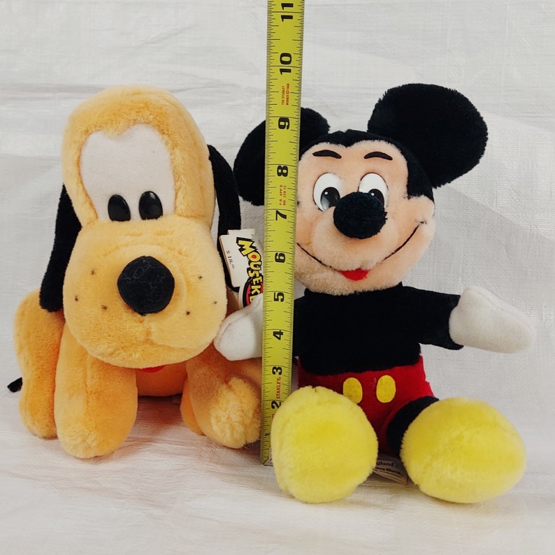 May include: Two plush toys, a yellow and brown dog and a black and red Mickey Mouse. The dog is sitting on the left and the Mickey Mouse is sitting on the right. The Mickey Mouse is holding a tag that says "Mouseke" in red letters.