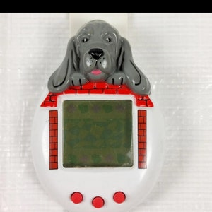 Puede incluir: Un juguete digital para mascotas blanco y rojo con forma de casa con un perro gris asomándose por el techo. El perro tiene la lengua rosa y los ojos negros. El juguete tiene tres botones rojos en la parte inferior.