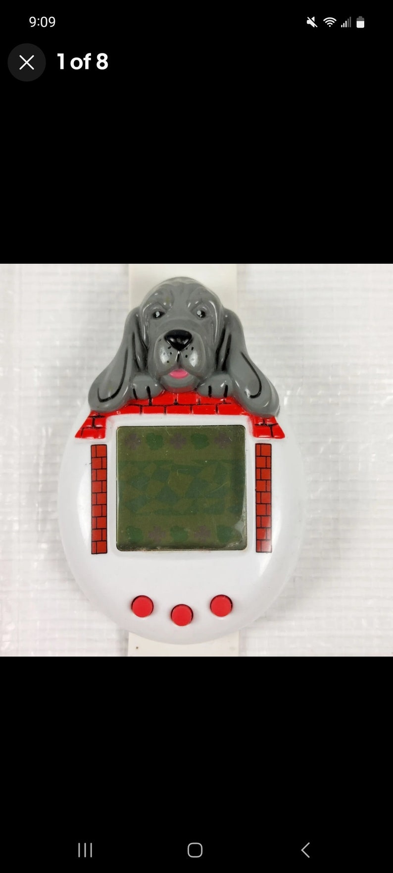 K&ouml;nnte beinhalten: Ein wei&szlig;es und rotes digitales Haustier-Spielzeug in Form eines Hauses mit einem grauen Hund, der &uuml;ber das Dach schaut. Der Hund hat eine rosa Zunge und schwarze Augen. Das Spielzeug hat drei rote Kn&ouml;pfe unten.