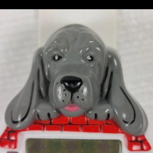 K&ouml;nnte beinhalten: Eine graue Plastik-Hundefigur mit rosa Zunge, die &uuml;ber einem roten Backsteinhaus mit einem wei&szlig;en digitalen Display lugt.