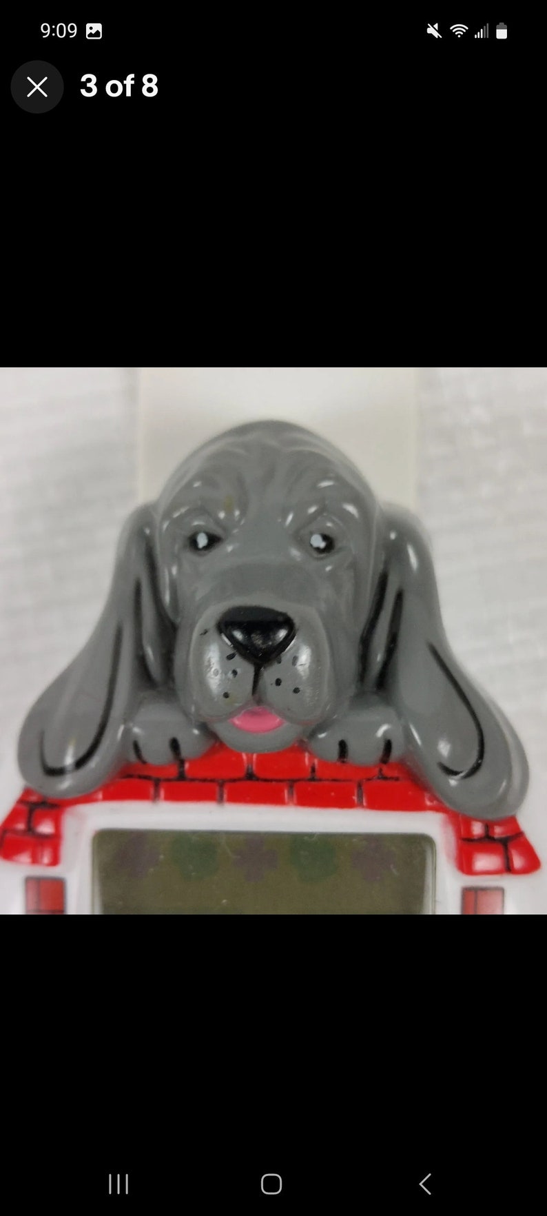 K&ouml;nnte beinhalten: Eine graue Plastik-Hundefigur mit rosa Zunge, die &uuml;ber einem roten Backsteinhaus mit einem wei&szlig;en digitalen Display lugt.