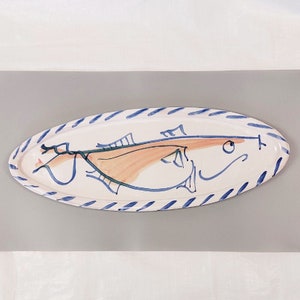 Op de afbeelding: Witte ovale keramische serveerschaal met een blauw-witte gestreepte rand. Een gestileerd visontwerp is in blauw, groen en oranje op de schaal geschilderd.