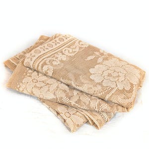May include: A folded beige lace fabric featuring a detailed floral pattern and a decorative border with geometric shapes. The textile has a semi-sheer, open-weave construction with intricate needlework that highlights the floral motifs throughout.