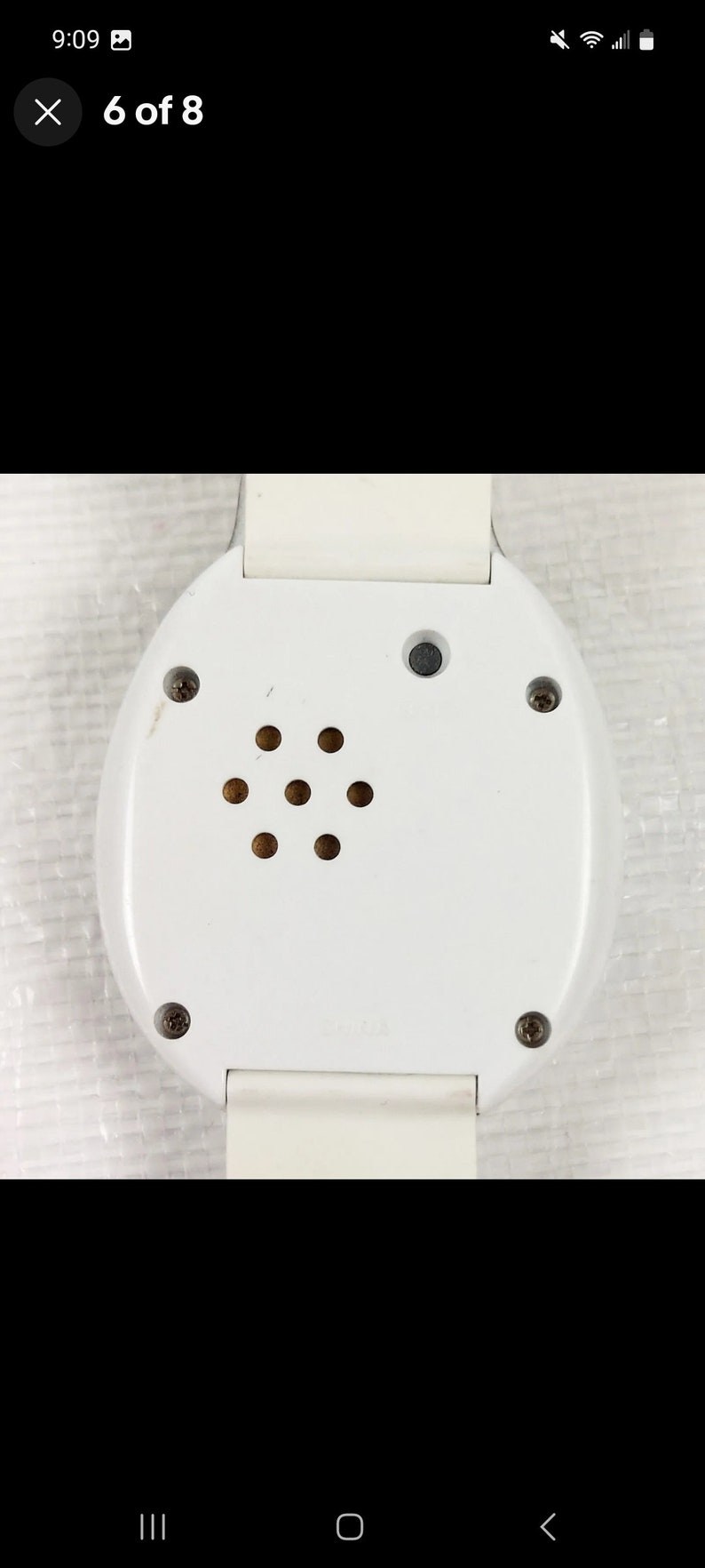 K&ouml;nnte beinhalten: Wei&szlig;e Armbanduhr mit einem runden Zifferblatt und einem kleinen schwarzen Knopf oben. Die Uhr hat eine silberne Schraube an der Seite und einen Lautsprecher mit mehreren L&ouml;chern unten.