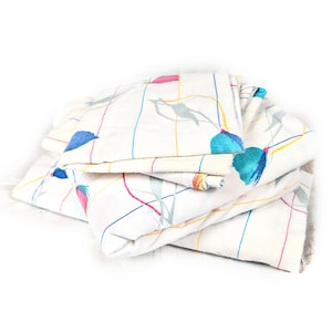May include: Folded white fabric with a floral pattern. The design features blue, pink, and gray floral elements, along with a grid of colorful lines. The fabric is folded in a stack, showcasing the pattern.