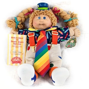 May include: A Cabbage Patch Kids Circus Kids doll with blonde pigtails, blue eyes, and a painted face. The doll wears a blue hat, suspenders, a rainbow tie, and a patterned shirt. It holds a birth certificate and adoption papers.