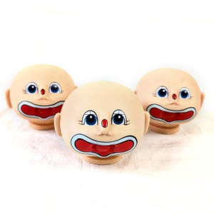 Pode incluir: Três cabeças de bonecas palhaço vintage com traços pintados. Cada cabeça tem um tom de pele claro, olhos azuis, um nariz vermelho e uma boca grande vermelha com um contorno branco e azul. As cabeças estão dispostas sobre uma superfície branca.