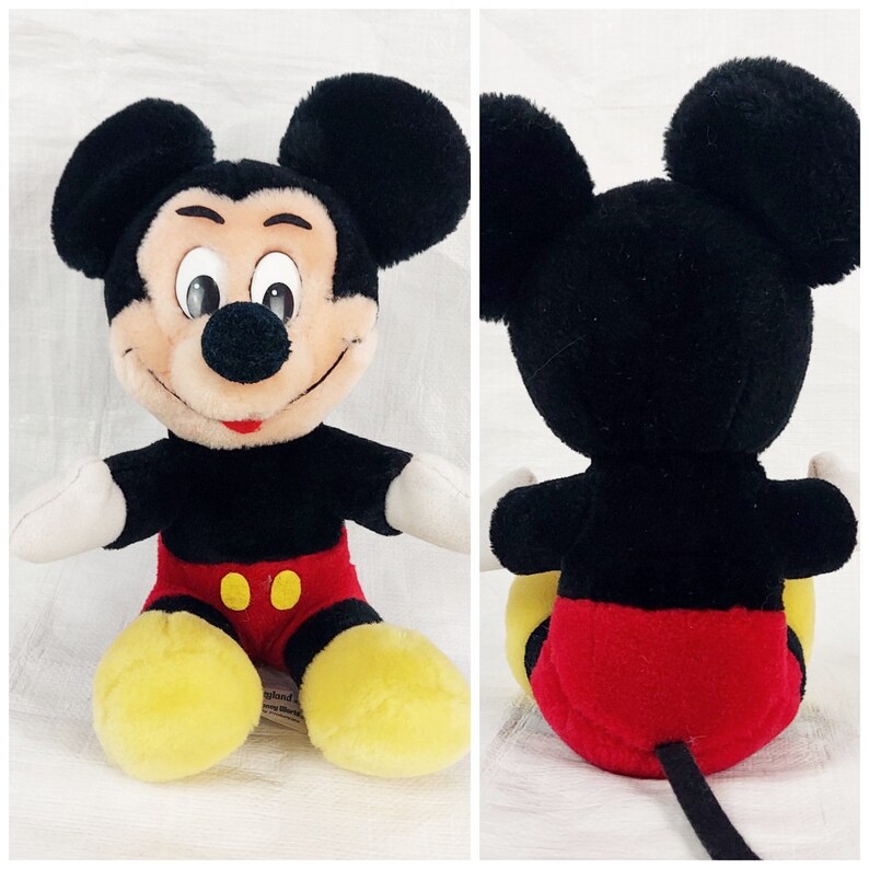 May include: A plush Mickey Mouse toy sitting with its legs crossed. The toy is black with a red and yellow outfit. The toy has a black tail.