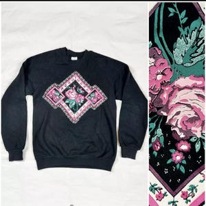 May include: Black sweatshirt with a diamond-shaped floral design featuring pink and green roses. The design is centered on the front of the sweatshirt.