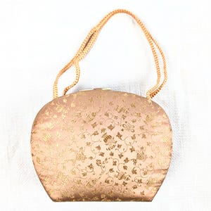 May include: A vintage, oval-shaped handbag in a soft peach color with a gold floral pattern. The bag has a matching peach-colored, braided handle. The clasp is a gold-colored metal. The bag is likely made of a satin-like material.