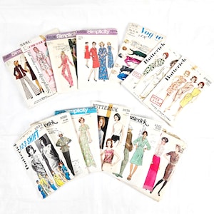 Puede incluir: Una colección de patrones de costura vintage, incluyendo marcas como Simplicity y Butterick. Los patrones presentan ilustraciones de vestidos y monos. Los patrones están dispuestos sobre una superficie blanca.