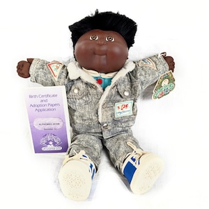 Könnte beinhalten: Cabbage Patch Kids Puppe, Alphonso Dean, trägt eine Jeansjacke und -hose mit weißen und blauen Turnschuhen. Die Puppe hat schwarzes Haar und einen dunklen Teint. Ein Geburtsurkunde und Adoptionspapiere sind enthalten.