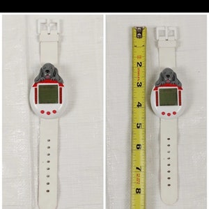 K&ouml;nnte beinhalten: Wei&szlig;e Armbanduhr aus Kunststoff mit einem rot-wei&szlig;en Digitaldisplay und einer grauen Hundgrafik. Die Uhr hat einen Durchmesser von 5 cm und ein wei&szlig;es Kunststoffband.