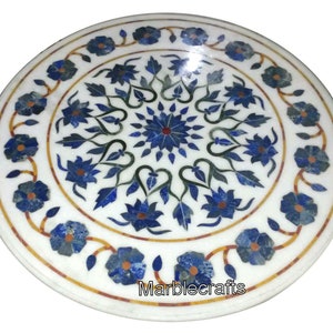 May include: Circular white marble table top with detailed floral inlay. The design incorporates blue and green floral patterns, with a central starburst. A repeating floral pattern with yellow and orange accents forms the border. The text "Marblecrafts" is visible.