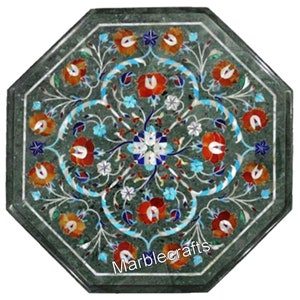 Könnte beinhalten: Eine achteckige Tischplatte aus grünem Marmor mit aufwendigen floralen Einlegearbeiten aus roten, orangen, weißen und blauen Steinen. Die Oberfläche zeigt ein symmetrisches Muster aus Blumen und Ranken, mit dem in Weiß gedruckten Text Marblecrafts.