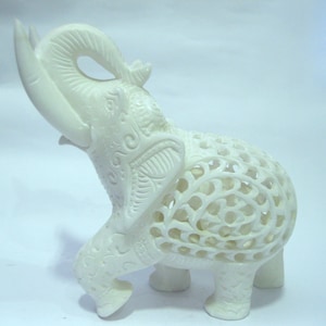 May include: Ivory-colored elephant figurine with intricate carved details. The elephant's trunk is raised, and its body features openwork patterns. A decorative piece for home decor.