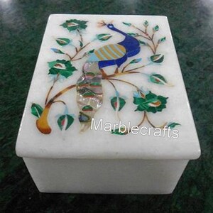 May include: A white marble box with a rectangular shape. The lid features a colorful peacock design with blue, yellow, and green accents, along with floral and leaf details. The text "Marblecrafts" is visible on the lid.