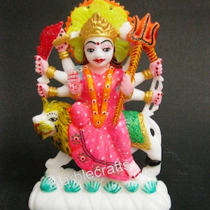 May include: A colorful statue of a deity, featuring multiple arms and a serene expression. The figure is adorned in pink and gold attire, riding a lion. The base is white with green accents, and the text "Marblecrafts" is visible.