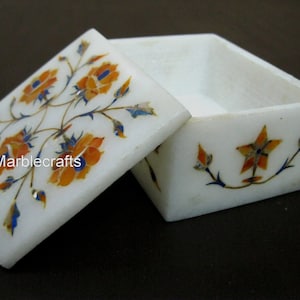 Può includere: Scatola in marmo bianco con coperchio, con intarsi floreali arancioni, blu e gialli. La scatola è aperta, rivelando l'interno. Il testo "Marblecrafts" è visibile sul coperchio. Contenitore decorativo.