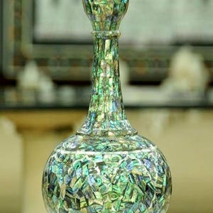 Puede incluir: Un jarrón decorativo con un cuerpo bulboso y un cuello delgado, adornado con un mosaico de piezas iridiscentes y multicolores. El jarrón tiene una parte superior acampanada y una base de pedestal, mostrando una mezcla de tonos verdes, azules y dorados.