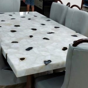 May include: A rectangular dining table with a white, semi-translucent stone surface embedded with dark, irregular stone accents. The table is surrounded by six light gray upholstered chairs, each featuring a curved, dark brown wooden handle.