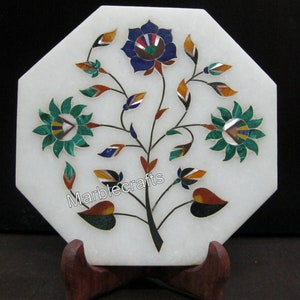Può includere: Pezzo decorativo ottagonale in marmo bianco con design floreale intarsiato. Il design presenta un albero centrale con foglie e fiori colorati nelle tonalità del blu, verde, arancione e marrone. Il pezzo è supportato da un supporto in legno e il testo "Marblecrafts" è visibile.