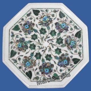 Puede incluir: Mesa octogonal de mármol blanco con diseño floral incrustado. La mesa presenta un patrón detallado de flores, hojas y tallos en tonos verdes, azules y materiales iridiscentes. La mesa mide aproximadamente 61 cm de diámetro.