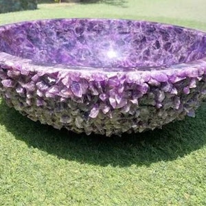 Può includere: Un lavabo rotondo in ametista viola con un esterno ruvido e strutturato. Il lavabo è appoggiato su erba verde, mettendo in risalto la sua bellezza naturale e il design unico. L'interno del lavabo è liscio e lucido.