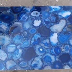 May include: A rectangular slab featuring a mosaic of polished blue agate slices. The surface displays a variety of deep navy, royal blue, and pale sky blue tones, with translucent white and grey crystalline centres embedded within the stone pattern.