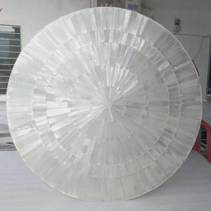 Puede incluir: Una gran tapa de mesa decorativa redonda y blanca. La superficie está hecha de muchas piezas pequeñas y rectangulares dispuestas en círculos concéntricos, que irradian desde el centro. La tapa de la mesa tiene un brillo nacarado.