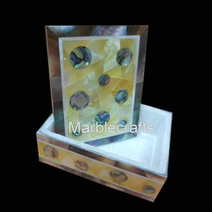 Op de afbeelding: Een decoratieve doos- en framesset. Het frame heeft een gele achtergrond met ingelegde iriserende cirkels. De doos heeft een bijpassend ontwerp met de tekst "Marblecrafts". De set is gemaakt van lichtgekleurd materiaal.