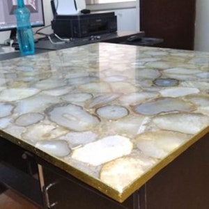 May include: A square desk featuring a tabletop composed of numerous polished, translucent white and beige agate slices embedded in a smooth, glossy resin surface, finished with a thin, metallic gold-toned edge trim on the perimeter.