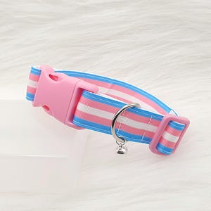 May include: A cat collar with a pink plastic buckle and a small silver bell. The collar has a design with blue, pink, and white stripes. The collar is designed with a pink and blue striped pattern.