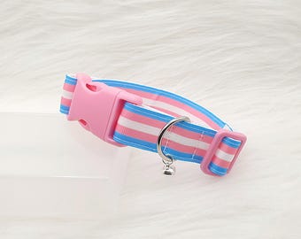 以下が含まれることがあります： ピンクとブルーのストライプ柄の猫用首輪。ピンク色のプラスチックバックルと小さなシルバーのベルが付いています。首輪は青、ピンク、白のストライプ模様のデザインです。