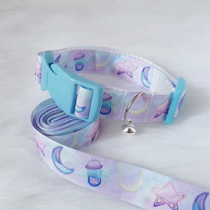 May include: A light blue adjustable cat collar with a bell and a matching purple and white ribbon with a moon and star pattern.