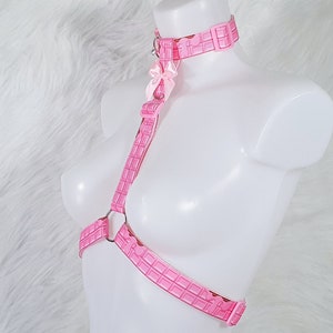 May include: Pink harness with a repeating chocolate bar pattern. The harness includes a choker, straps, and a chest piece, all connected with silver metal rings. A small pink bow adorns the front. The harness is displayed on a white mannequin against a white, fluffy background.