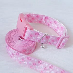 May include: Pink pet collar and leash set. The collar and leash are adorned with a repeating pattern of white paw prints. The collar has a pink plastic buckle and a small silver bell charm. The leash is rolled up, ready for use.