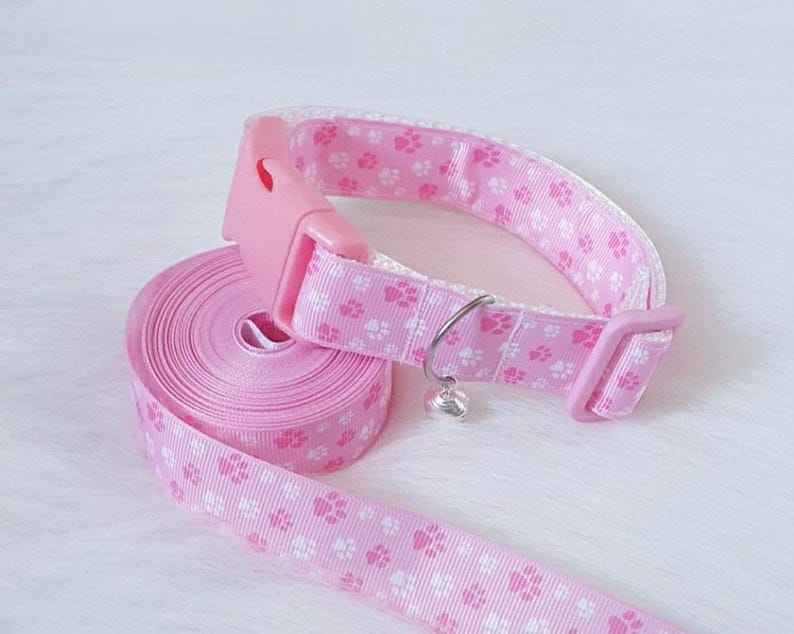 May include: Pink pet collar and leash set. The collar and leash are adorned with a repeating pattern of white paw prints. The collar has a pink plastic buckle and a small silver bell charm. The leash is rolled up, ready for use.
