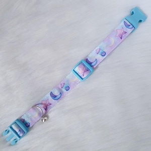 May include: A light blue adjustable collar with a white and purple pattern of moons and stars. The collar has a silver bell attached.