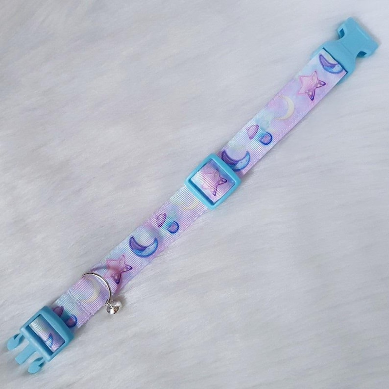 May include: A light blue adjustable collar with a white and purple pattern of moons and stars. The collar has a silver bell attached.