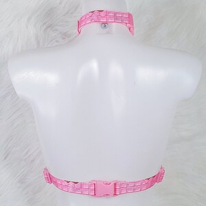 May include: Pink harness set with a grid pattern. The set includes a choker-style collar and a chest harness with a buckle closure. The harness is displayed on a white mannequin against a white, fluffy background.