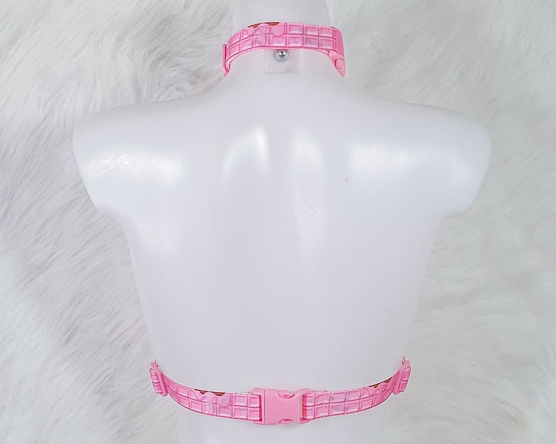 May include: Pink harness set with a grid pattern. The set includes a choker-style collar and a chest harness with a buckle closure. The harness is displayed on a white mannequin against a white, fluffy background.