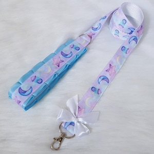 May include: A light blue and purple lanyard with a white bow and a silver key chain. The lanyard has a repeating pattern of moons, stars, and clouds.