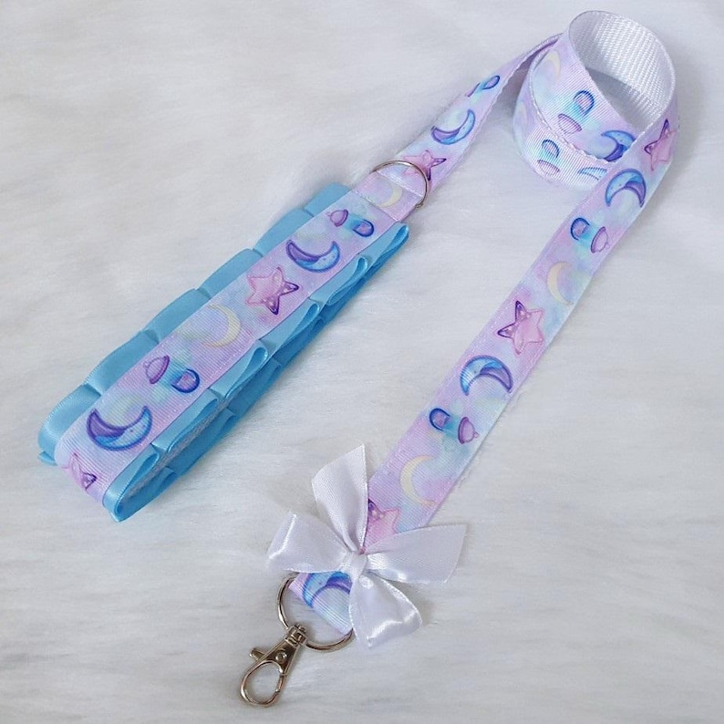 May include: A light blue and purple lanyard with a white bow and a silver key chain. The lanyard has a repeating pattern of moons, stars, and clouds.